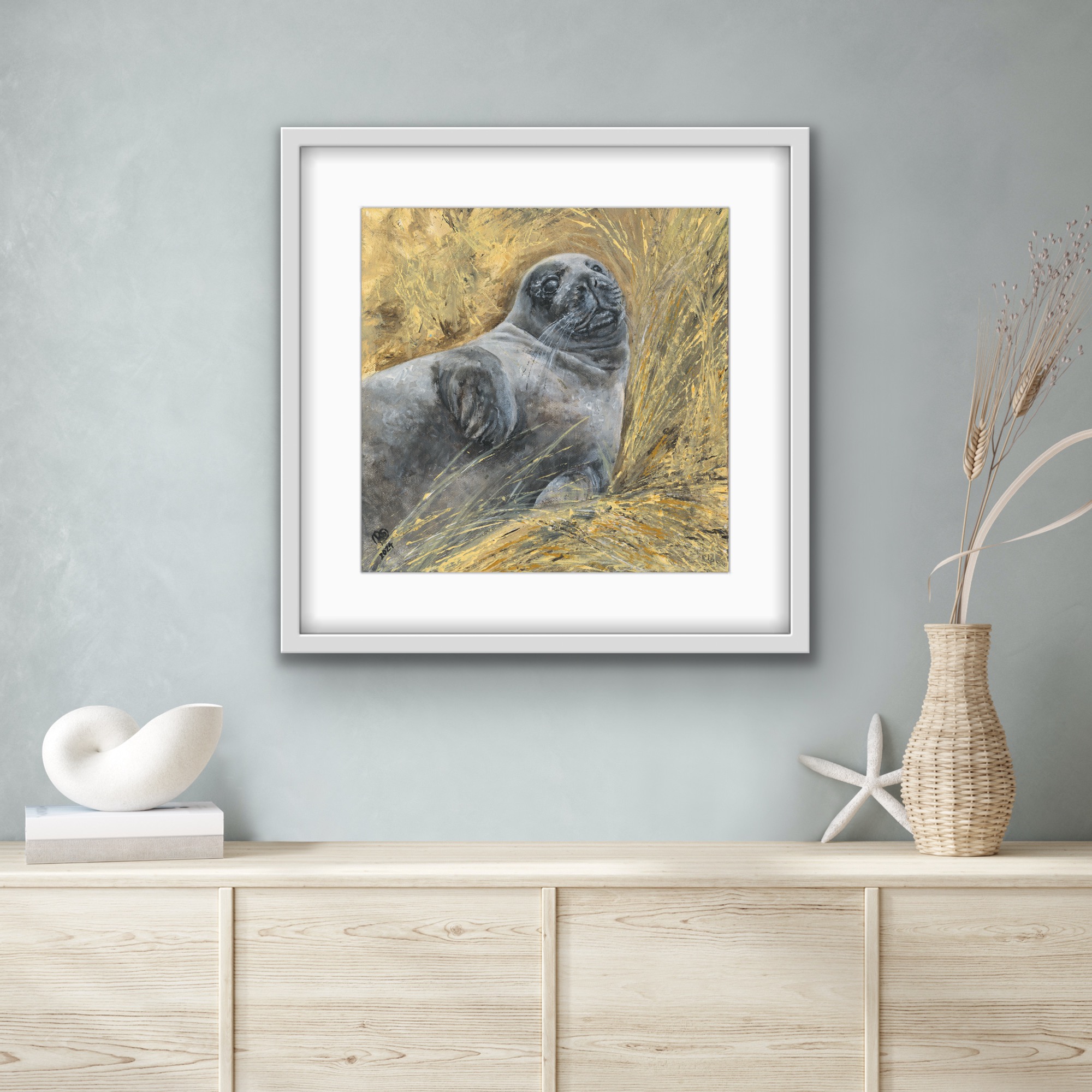 Held seal pup print in a pale grey frame with a white mount, styled above coastal accessories on a driftwood-toned surface